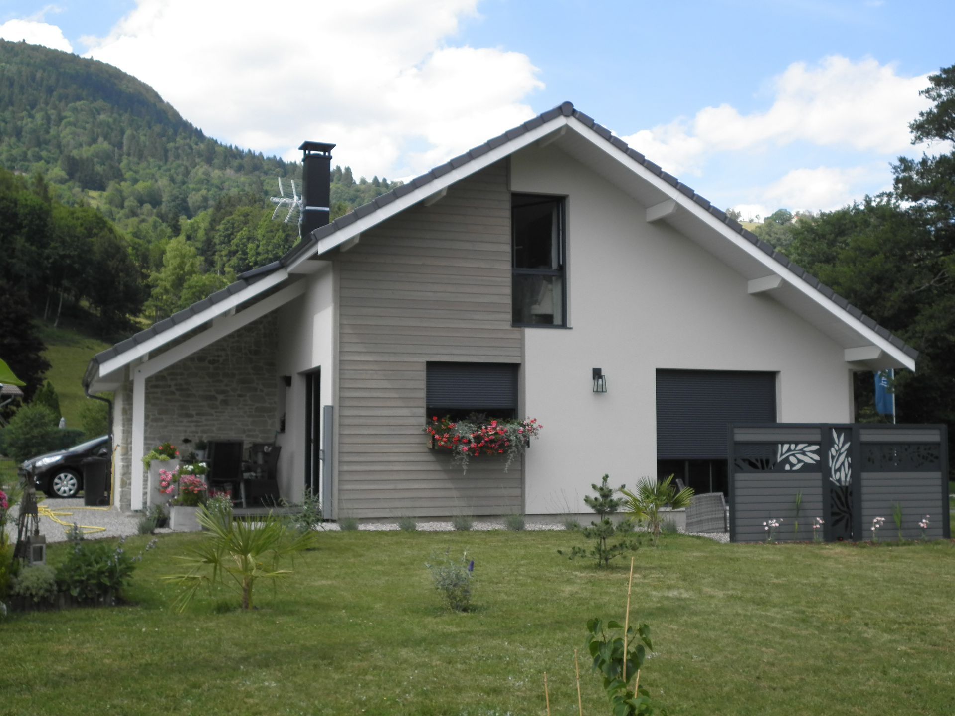 Maison grise moderne avec un toit pentu, une jardinière et un jardin verdoyant dans un cadre rural montagneux