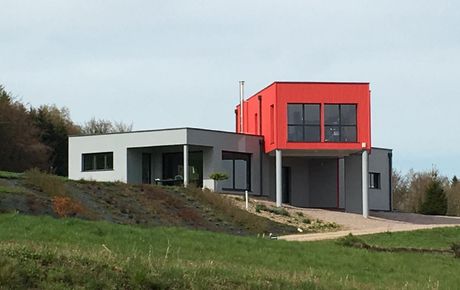 Maison moderne sur une colline herbeuse, avec un étage supérieur rouge et un niveau inférieur gris.