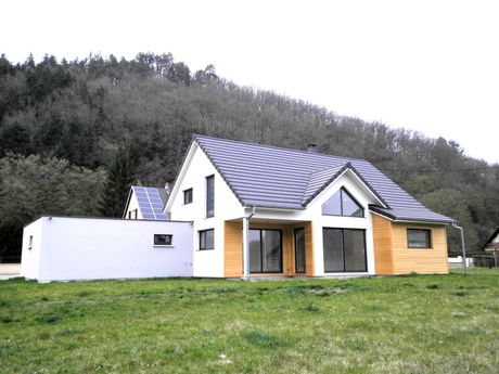 Maison moderne à l'extérieur blanc et en bois, située sur un coteau herbeux, adossée à des arbres.