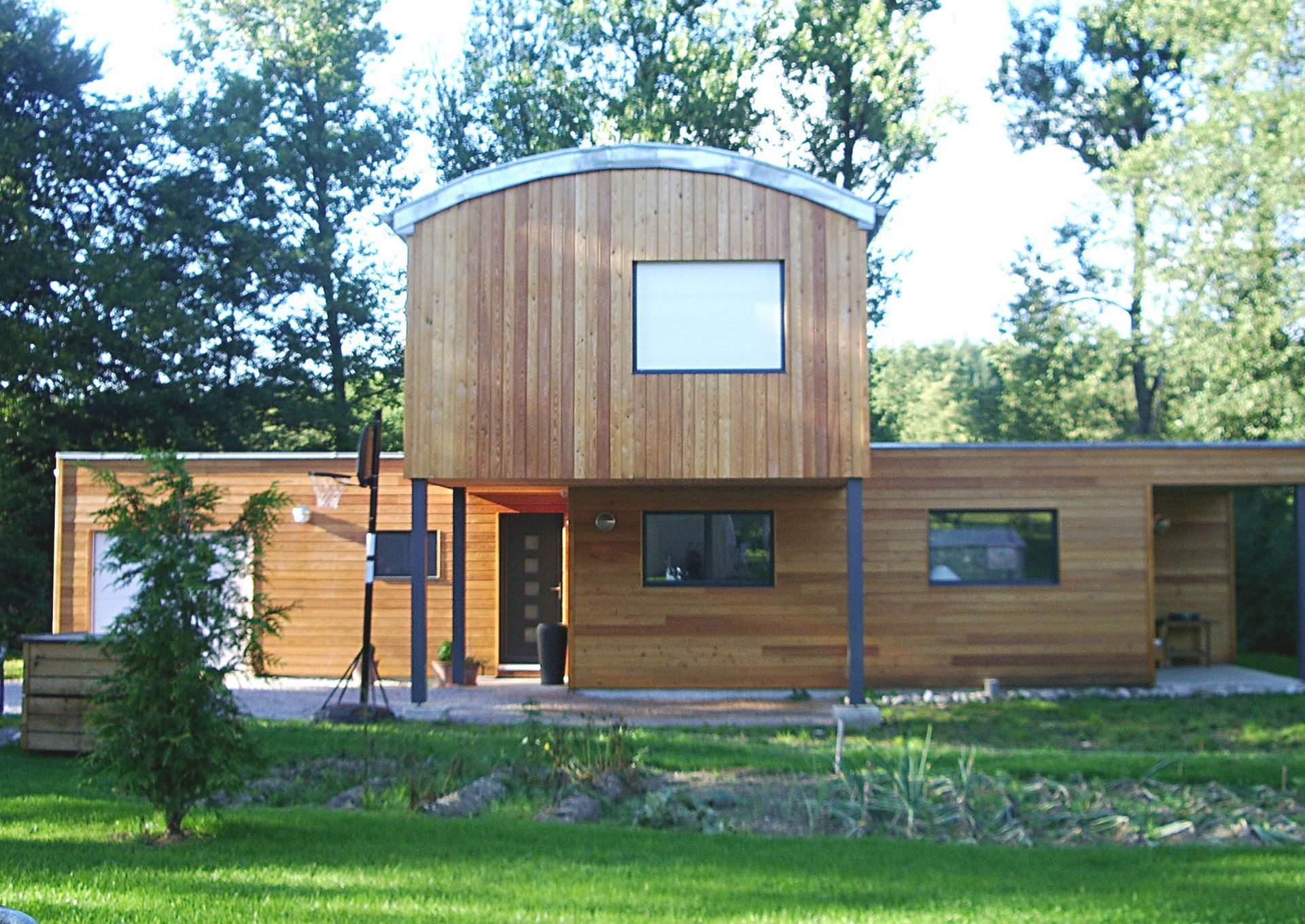 Maison moderne revêtue de bois avec une partie supérieure centrale incurvée, de grandes fenêtres et des arbres en façade