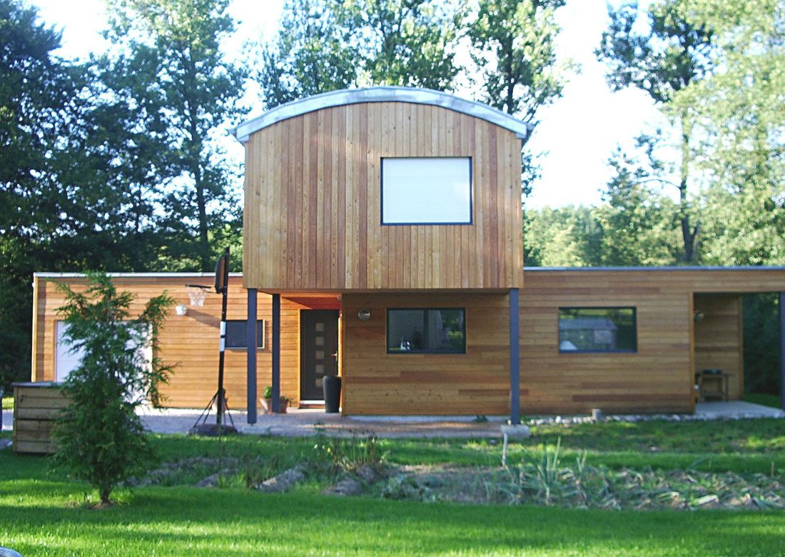 Maison moderne revêtue de bois avec une partie supérieure centrale incurvée, de grandes fenêtres et des arbres en façade