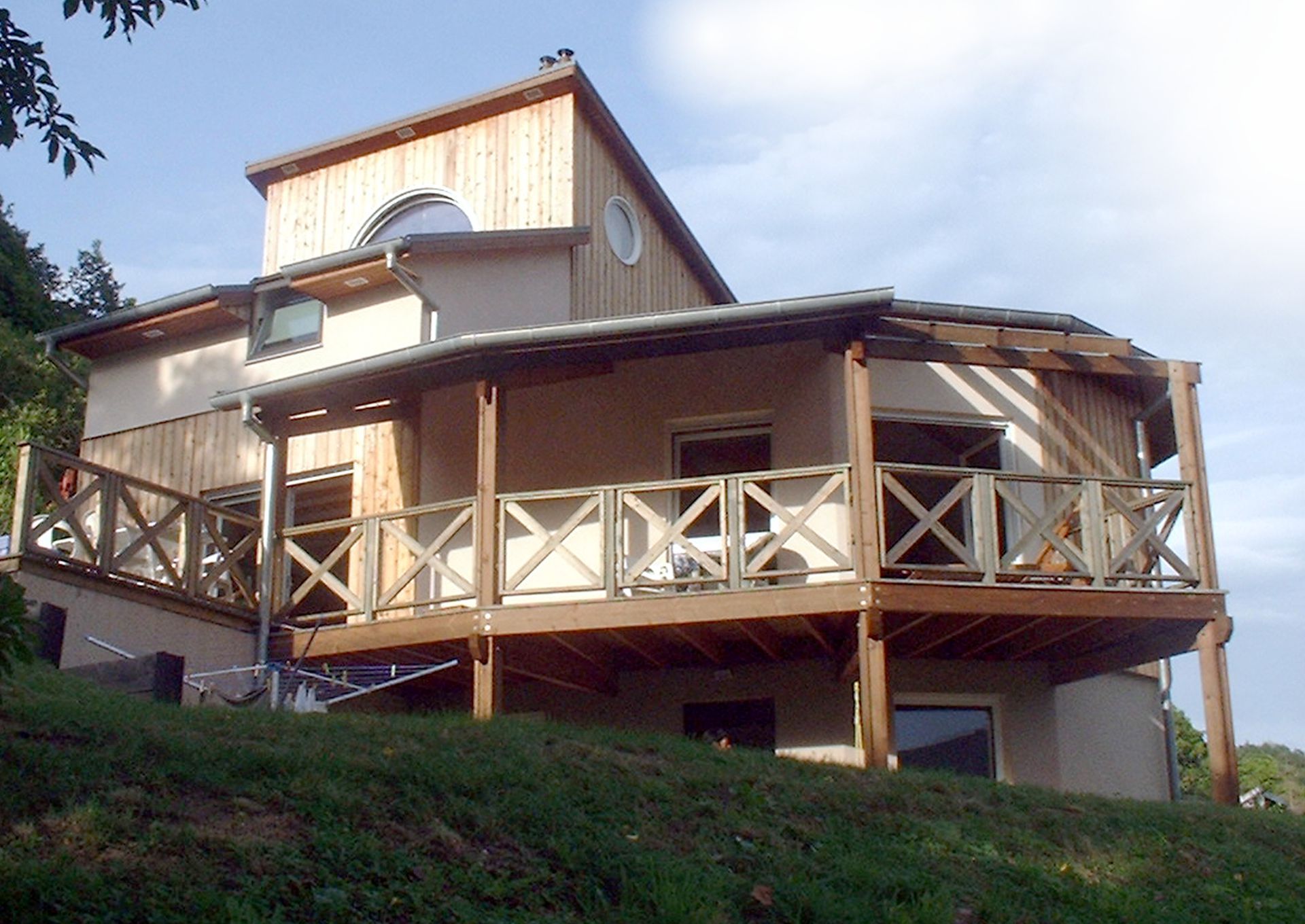 Maison beige à deux étages avec véranda en bois qui l'entoure, sur une pente herbeuse sous un ciel nuageux.