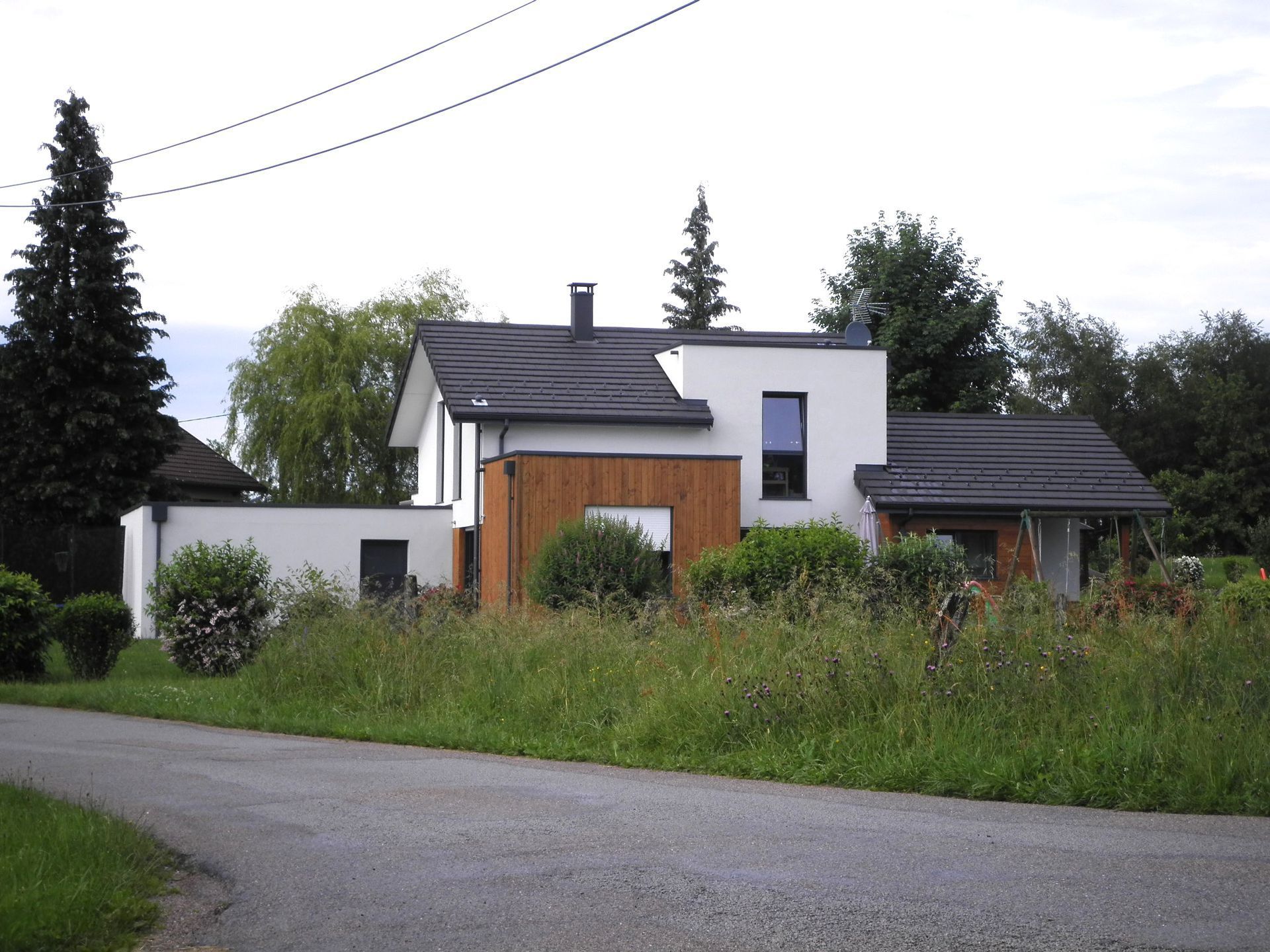 Maison blanche moderne avec des touches de bois, entourée d'arbres et de hautes herbes au bord d'une route sinueuse.