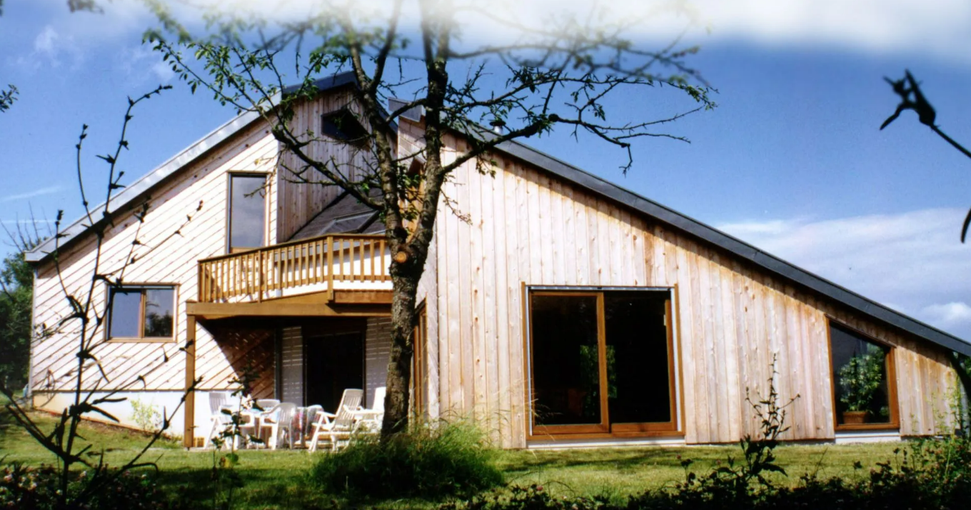 Maison moderne à deux étages avec bardage en bois clair, un petit balcon et de grandes fenêtres donnant sur un jardin gazonné.