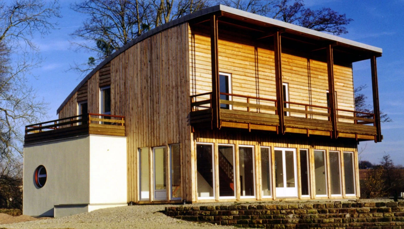 Maison moderne à deux étages avec bardage en bois, grandes fenêtres et balcon donnant sur un espace extérieur gazonné.