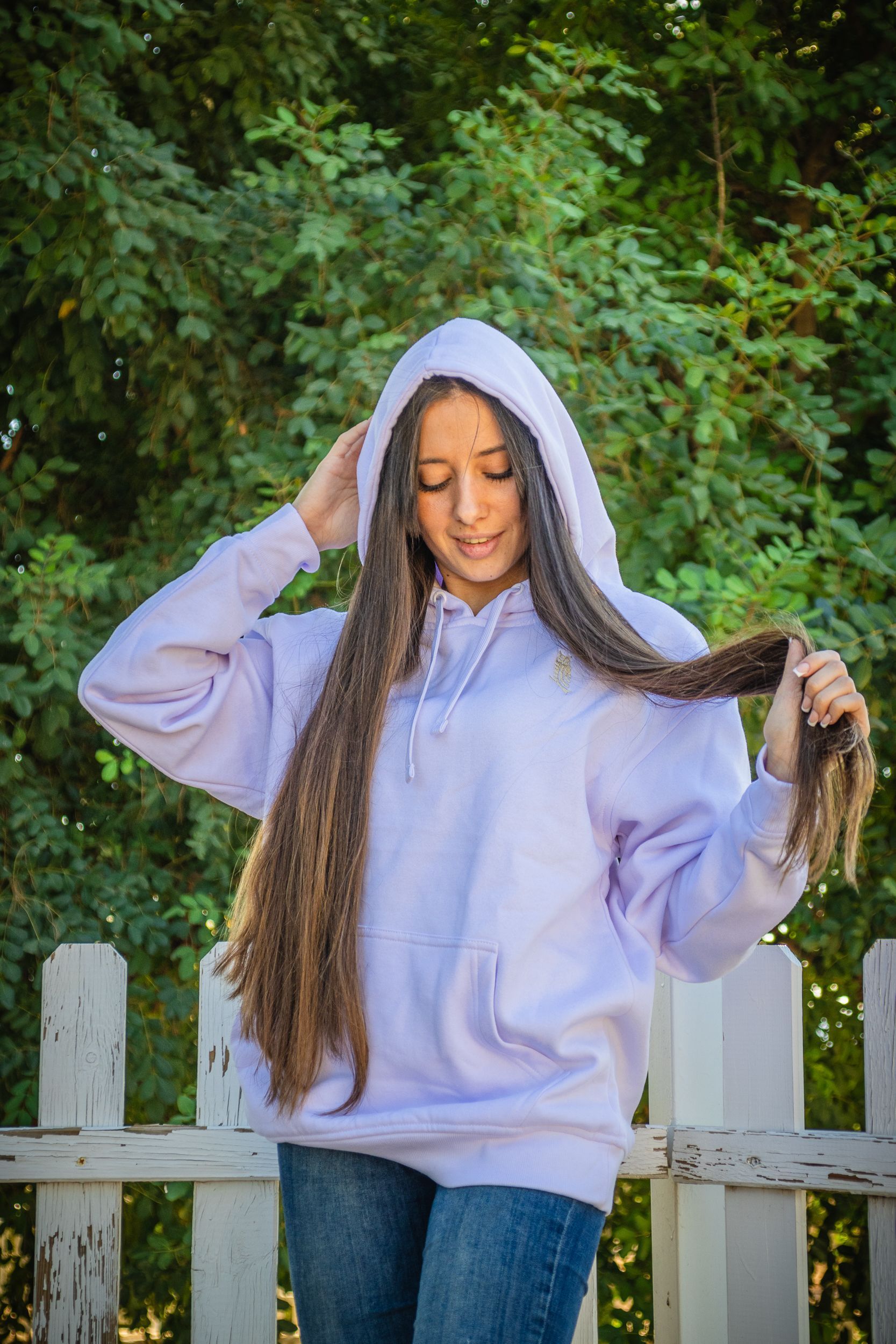 Una persona con una sudadera con capucha de color lila claro, sosteniendo trenzas, está de pie frente a una valla blanca y follaje verde.