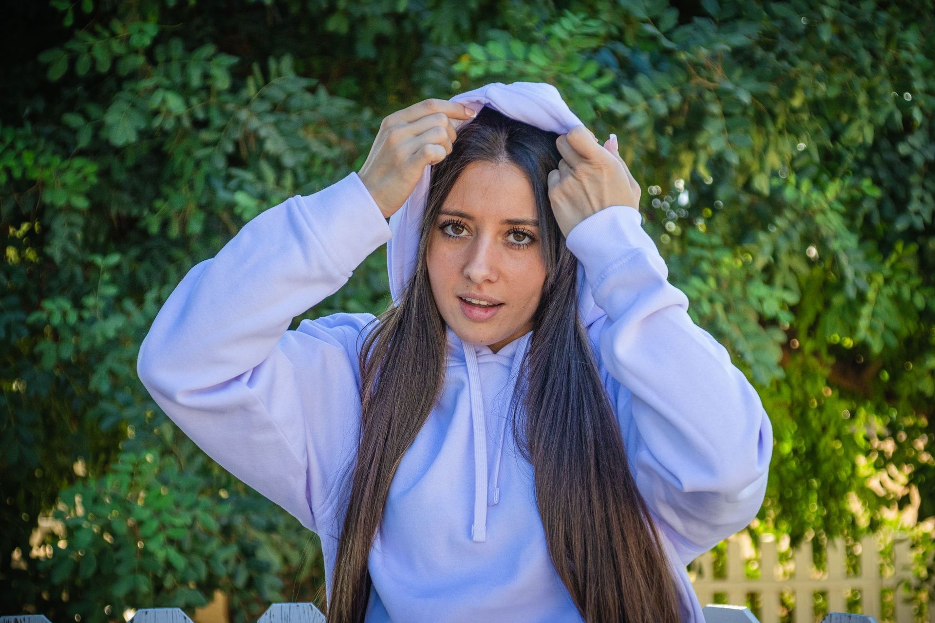 Una persona con una sudadera azul claro se ajusta la capucha al aire libre, frente a una vegetación verde.