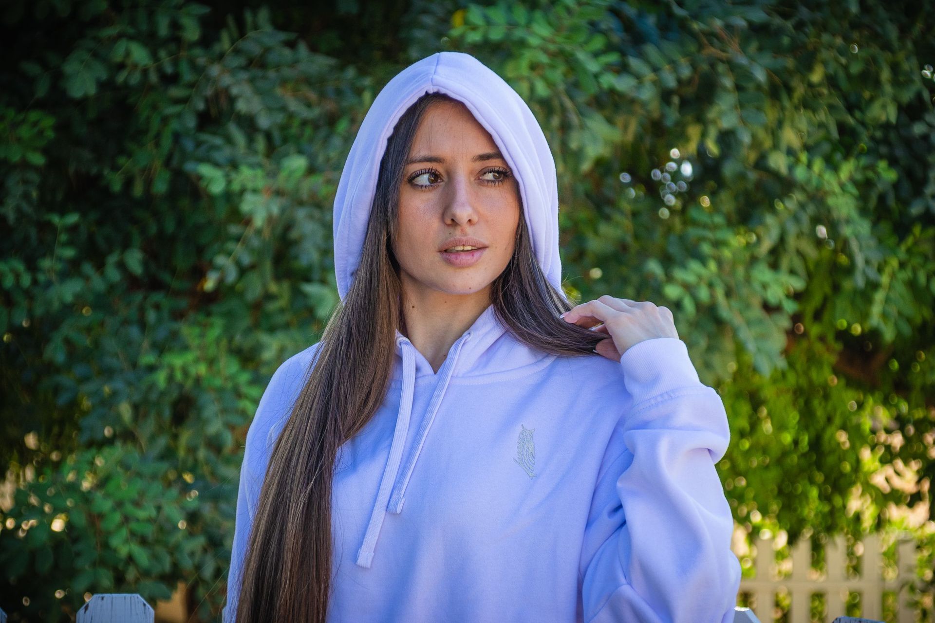 Persona con una sudadera blanca con capucha de pie al aire libre junto a la vegetación, mirando hacia un lado.