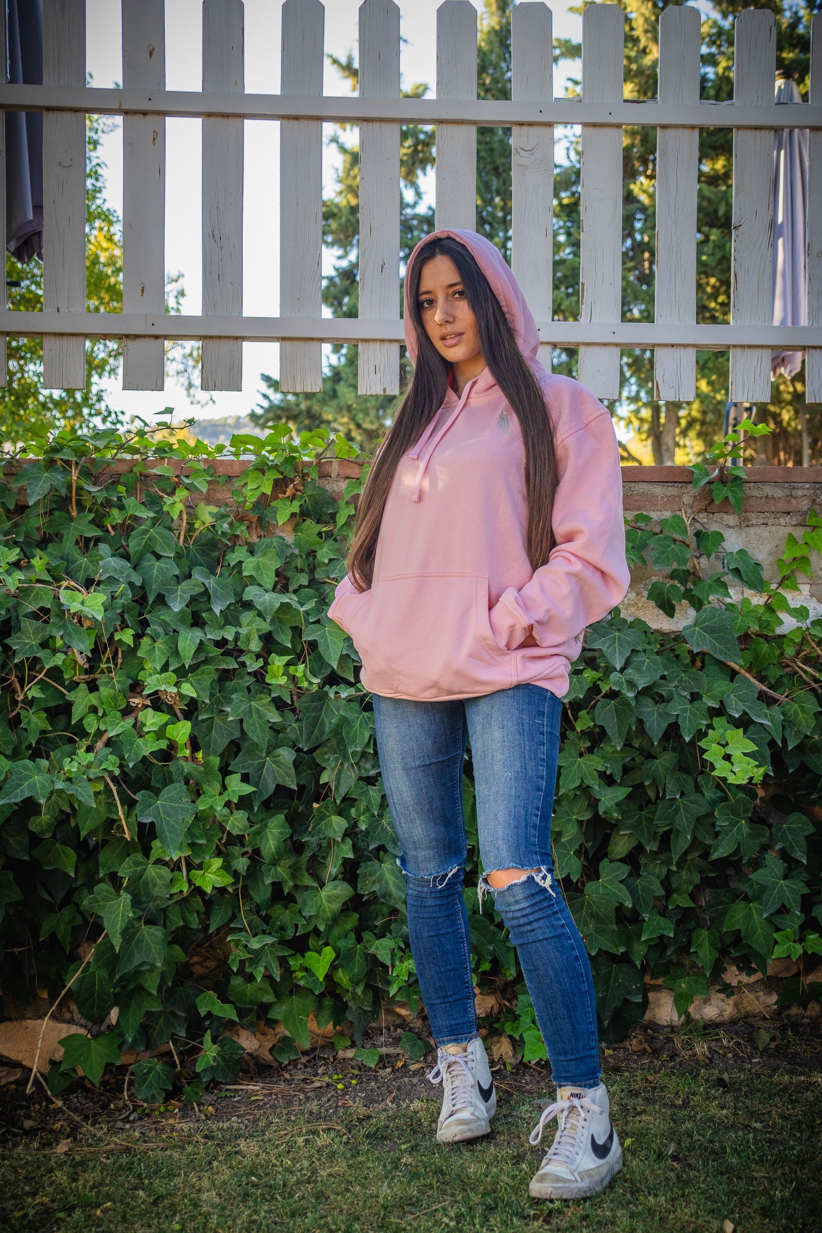Persona con sudadera rosa y vaqueros rotos de pie al aire libre junto a una valla blanca y vegetación.