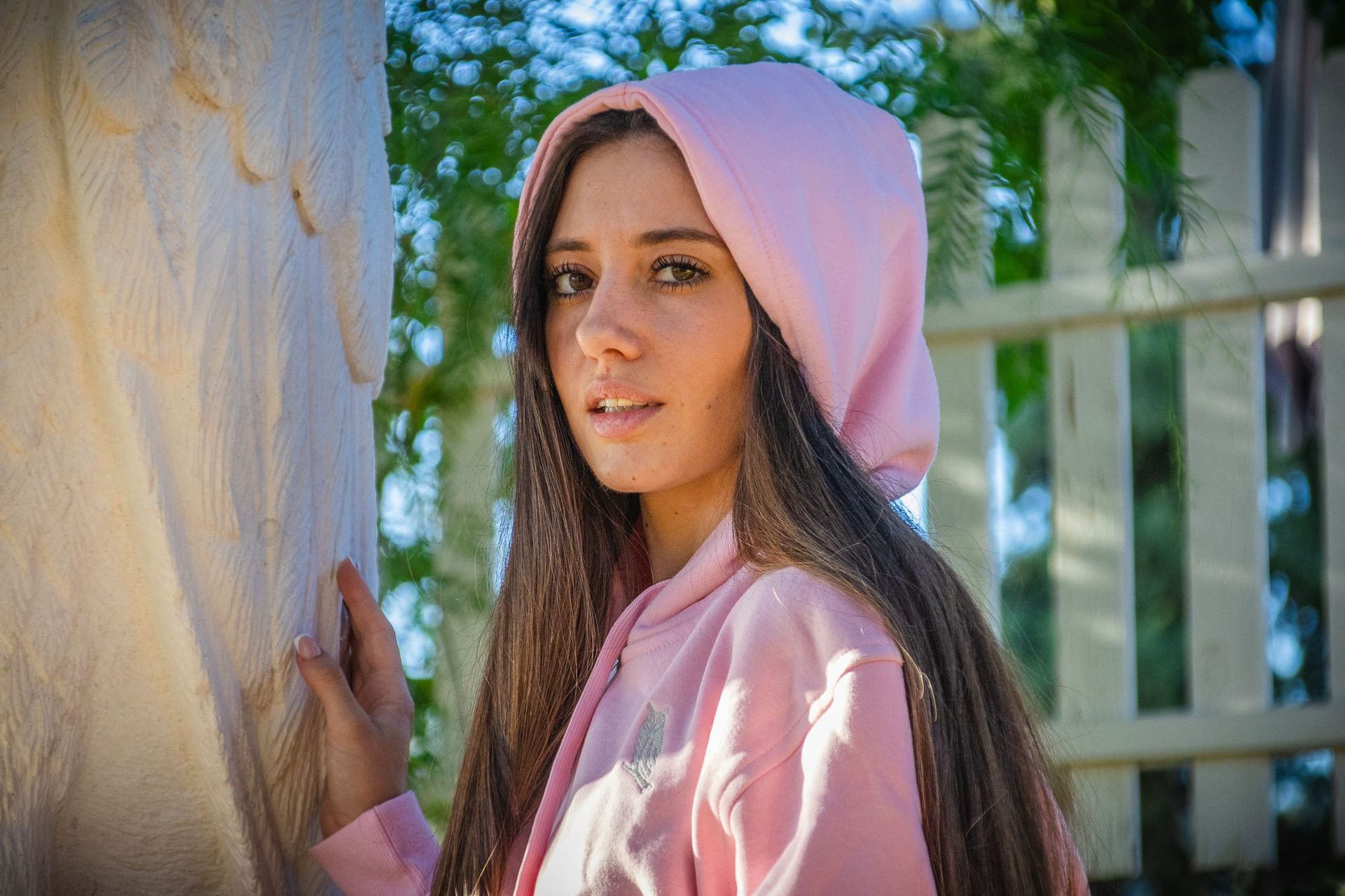 Mujer con sudadera rosa apoyada contra una pared al aire libre, mirando a la cámara.