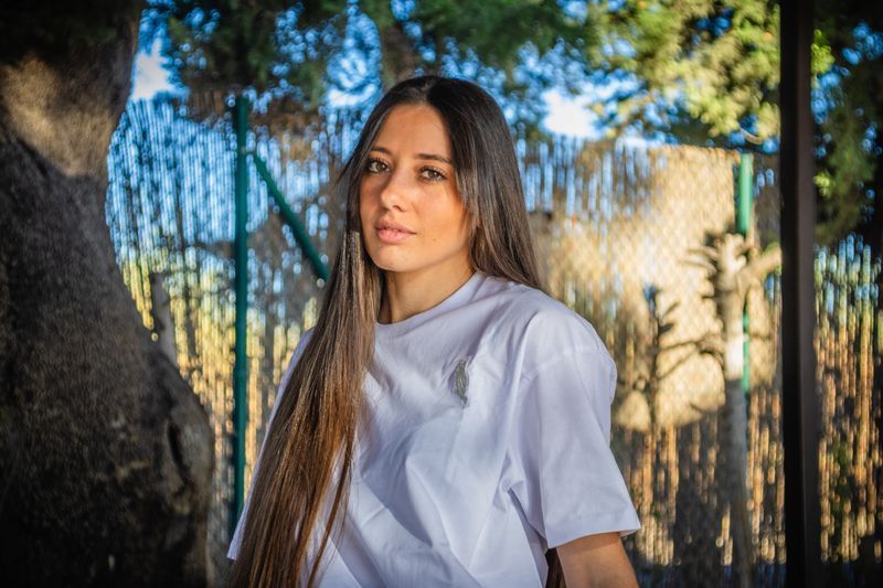 Mujer con camisa blanca de pie al aire libre cerca de un árbol y una cerca, con la luz del sol y vegetación borrosa detrás de ella.