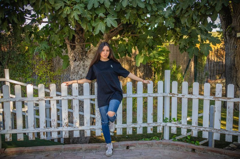 Mujer posando junto a una valla blanca bajo un árbol frondoso, vestida con una blusa negra y vaqueros rotos.