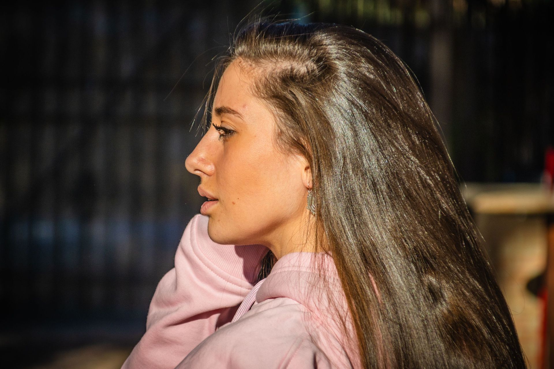 Perfil de una persona con cabello castaño largo, vestida con una sudadera rosa, al aire libre bajo la luz del sol.