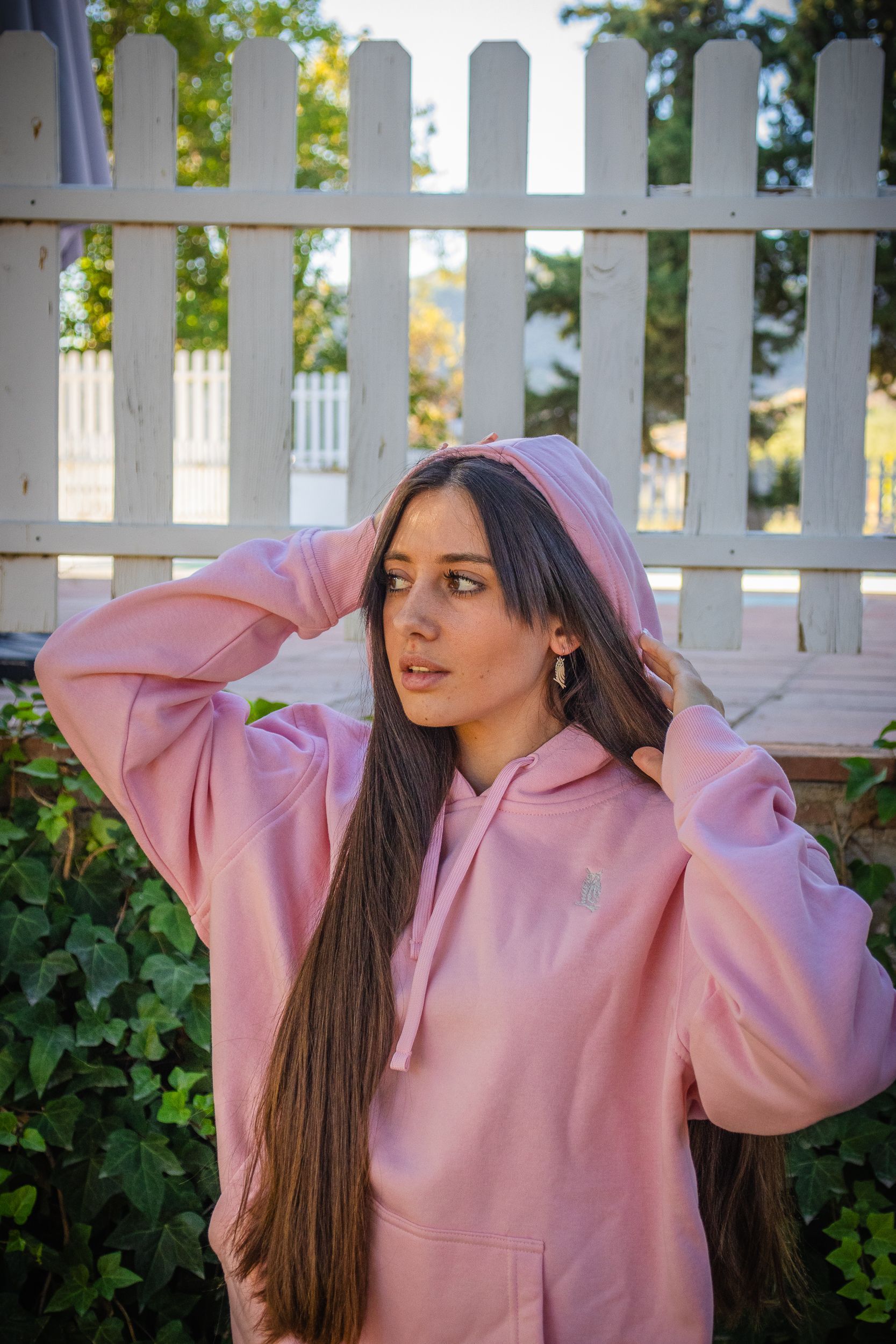 Persona con sudadera rosa y gorra morada posando junto a una valla blanca con vegetación detrás.