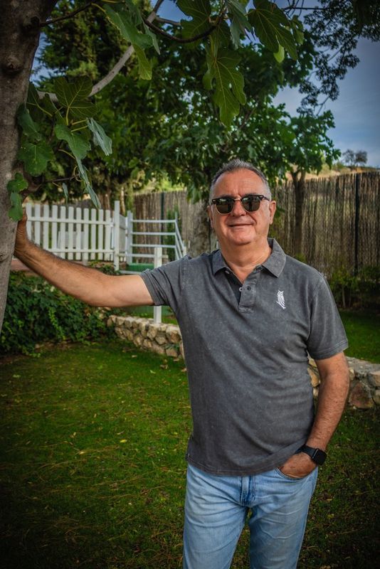 Hombre de pie al aire libre junto a un árbol y una valla blanca, con gafas de sol, polo gris y vaqueros, sonriendo.