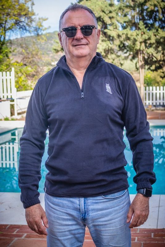 Hombre de pie junto a una piscina, con jersey azul marino, vaqueros y gafas de sol, con árboles y una valla al fondo.
