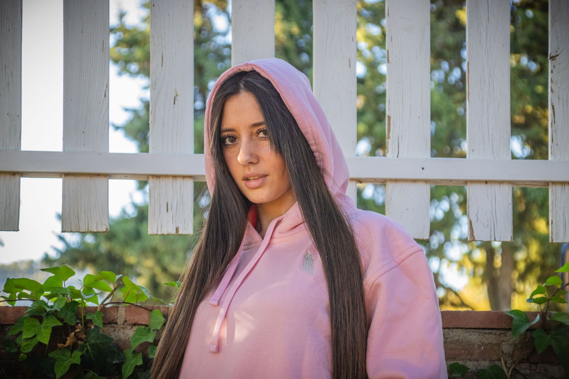 Mujer con sudadera rosa de pie junto a una valla blanca en un jardín soleado.