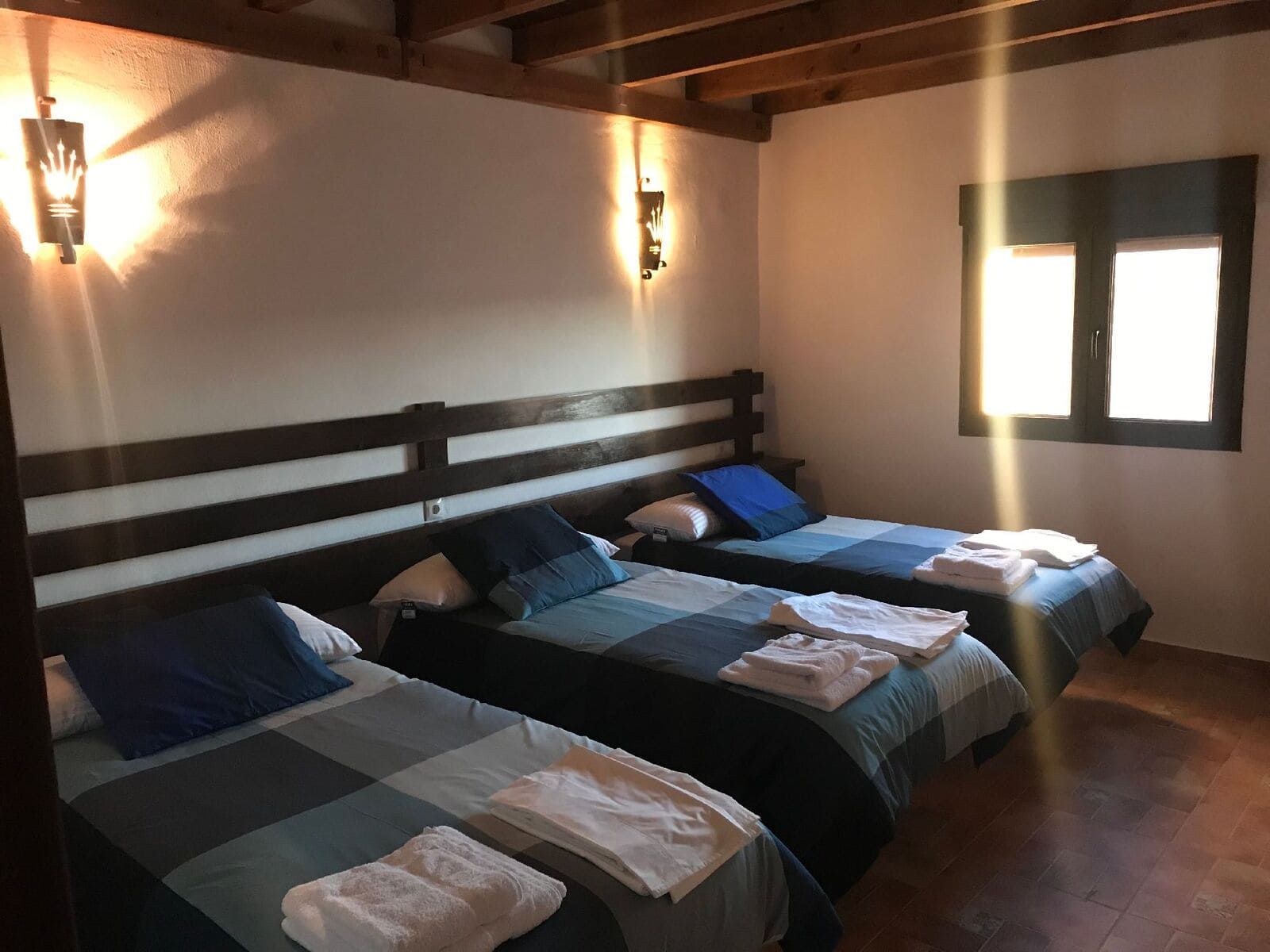 Tres camas en una habitación, cada una con una manta a cuadros azules y verdes y una toalla blanca. Luz del sol entrando por una ventana.