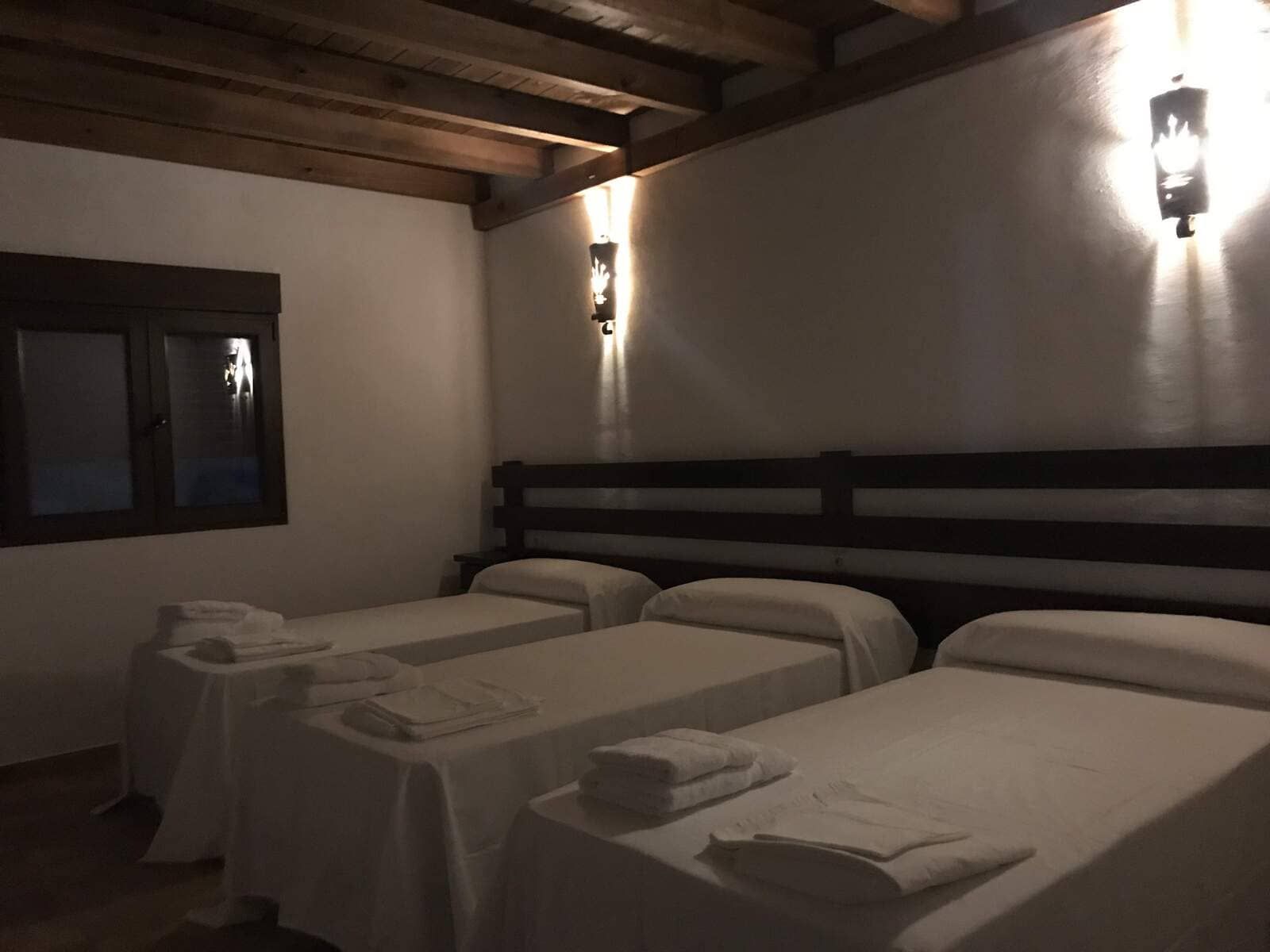 Dormitorio con tres camas individuales, sábanas blancas, vigas de madera e iluminación de aplique.