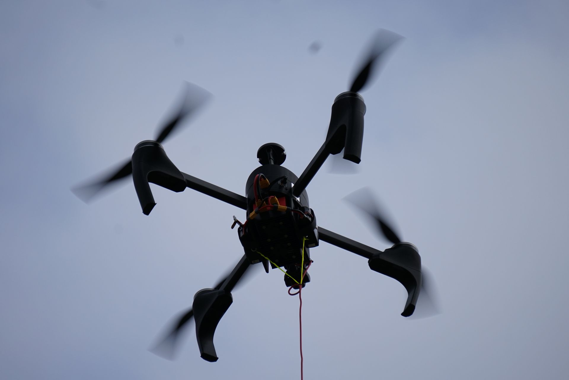 Eine schwarze Quadcopter-Drohne fliegt vor einem bewölkten Himmel und zieht einen dünnen roten Draht von ihrer Unterseite hinter sich her.
