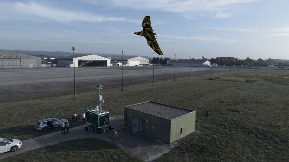 Eine getarnte Drohne fliegt über einem kleinen Gebäude und einer mobilen Kommandoeinheit auf einem Flugfeld.