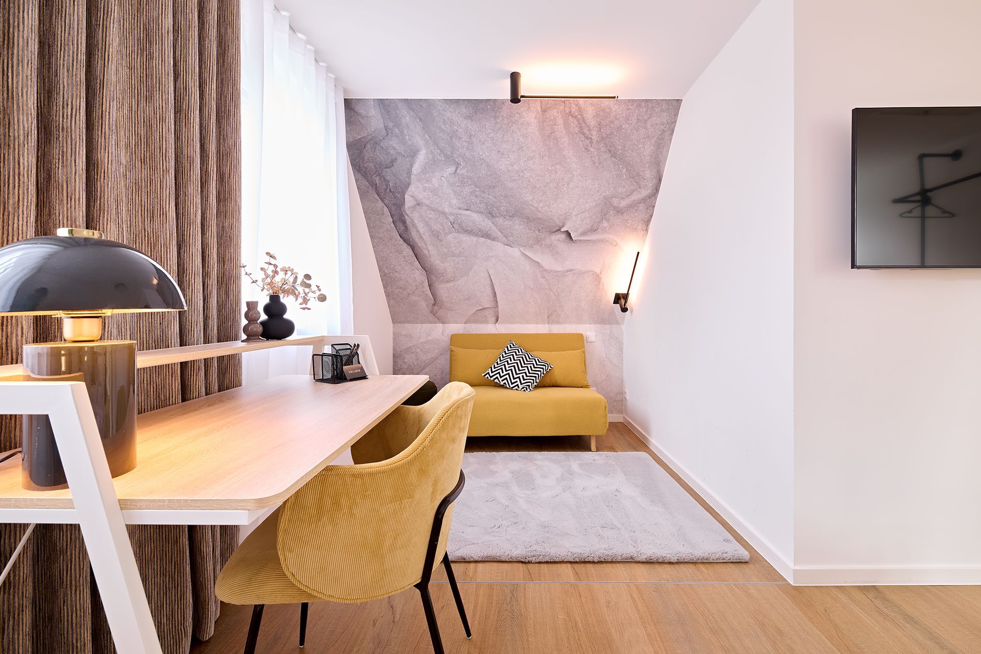 Ein Wohnzimmer mit einer gelben Couch, einem Schreibtisch, einem Sessel und einem Fernseher.
