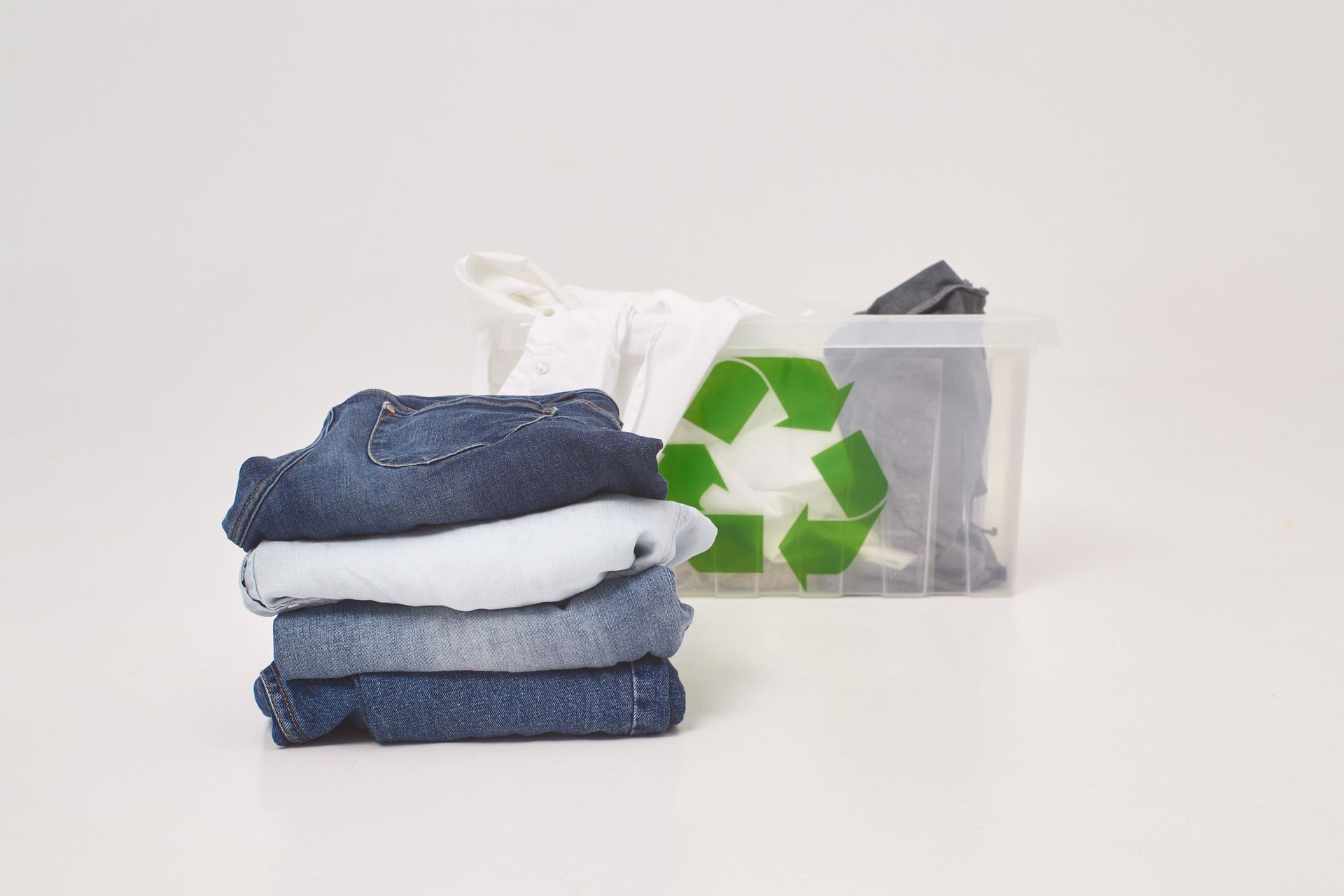 Pila de jeans doblados al lado de un contenedor con un símbolo de reciclaje y ropa.