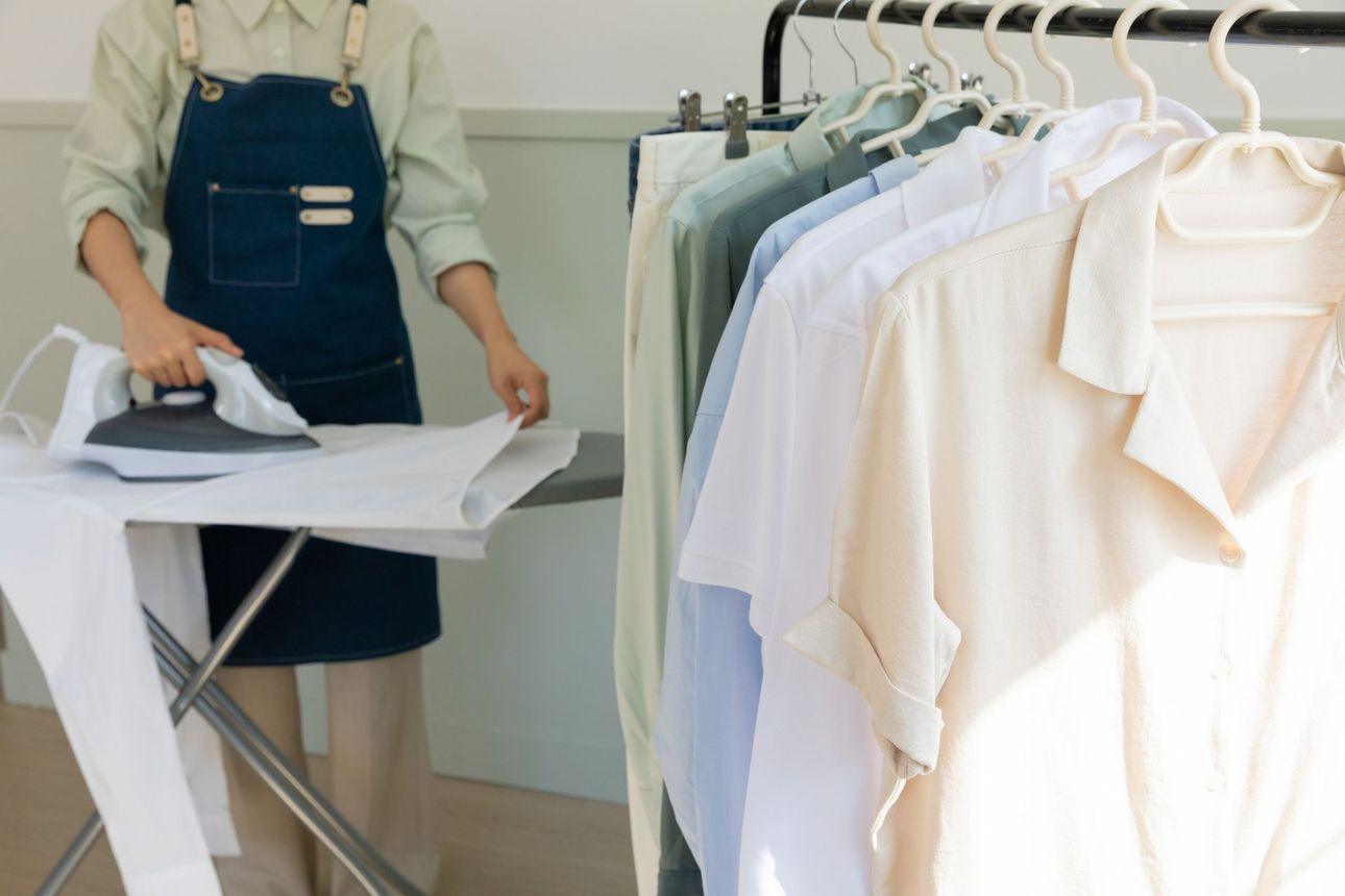 Persona planchando una camisa blanca sobre una tabla; camisas colgadas en un perchero cercano en una habitación de color claro.
