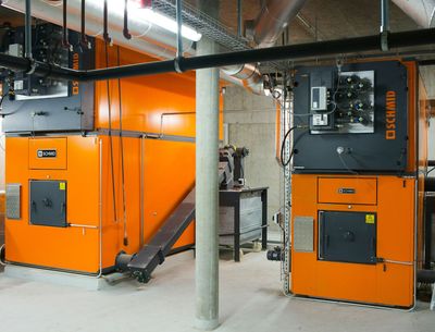 Deux chaudières industrielles orange dans une buanderie au sous-sol, avec des tuyaux apparents et des colonnes en béton.