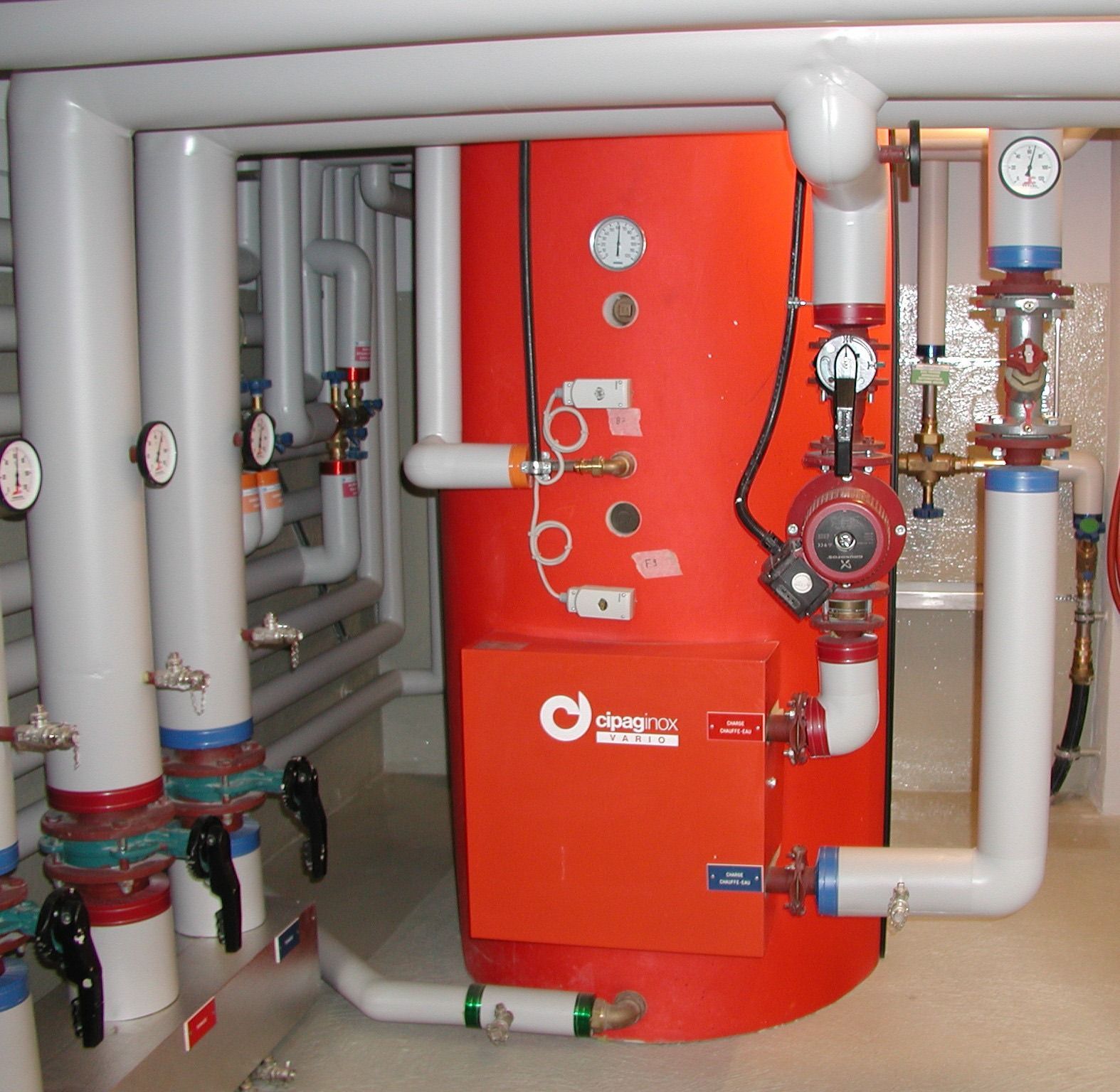 Chaufferie avec un grand réservoir d'eau chaude rouge, des tuyaux, des manomètres et des vannes.