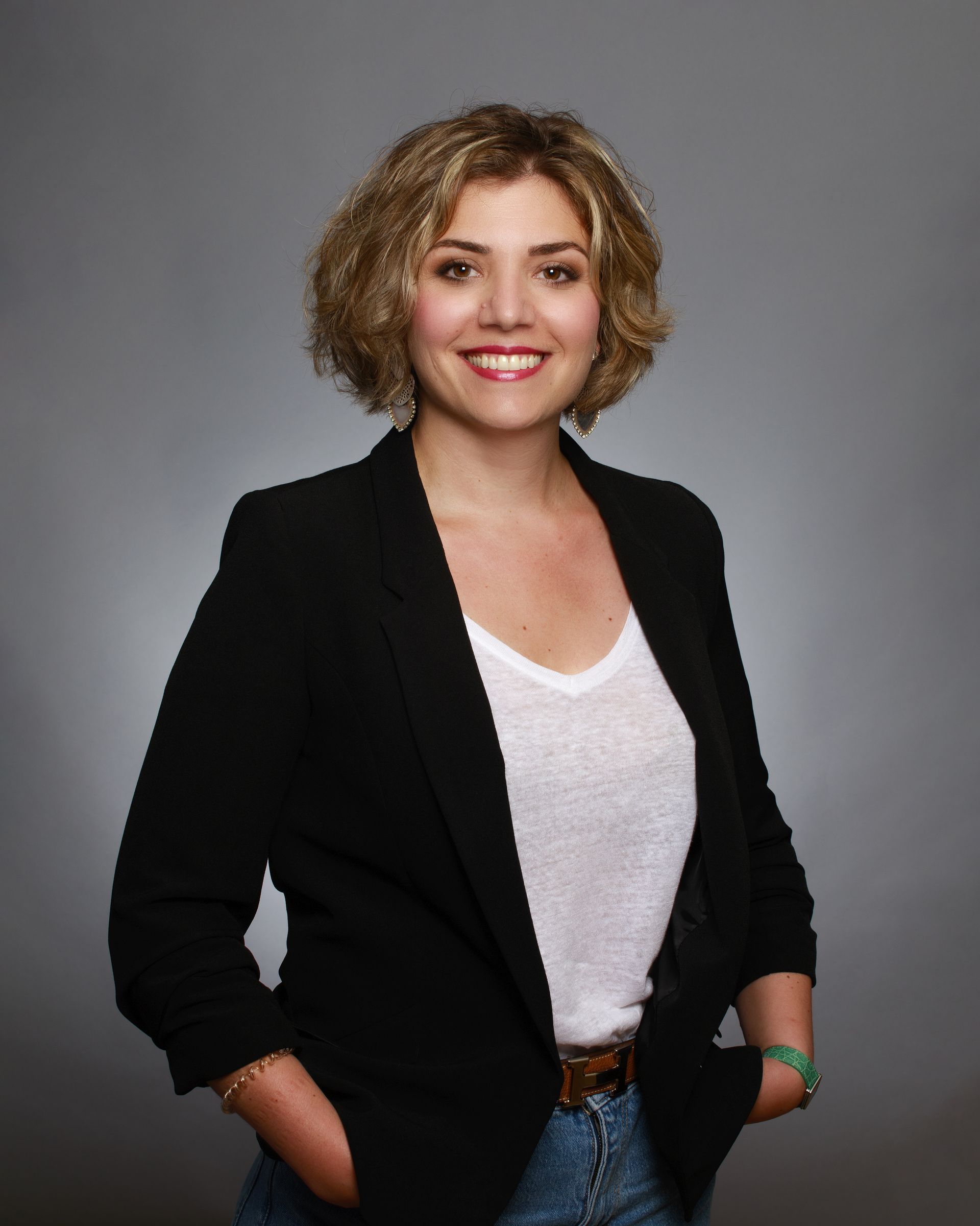 Une femme aux cheveux blonds courts et bouclés sourit, vêtue d'un blazer noir, d'un haut blanc et d'un jean, les mains dans les poches