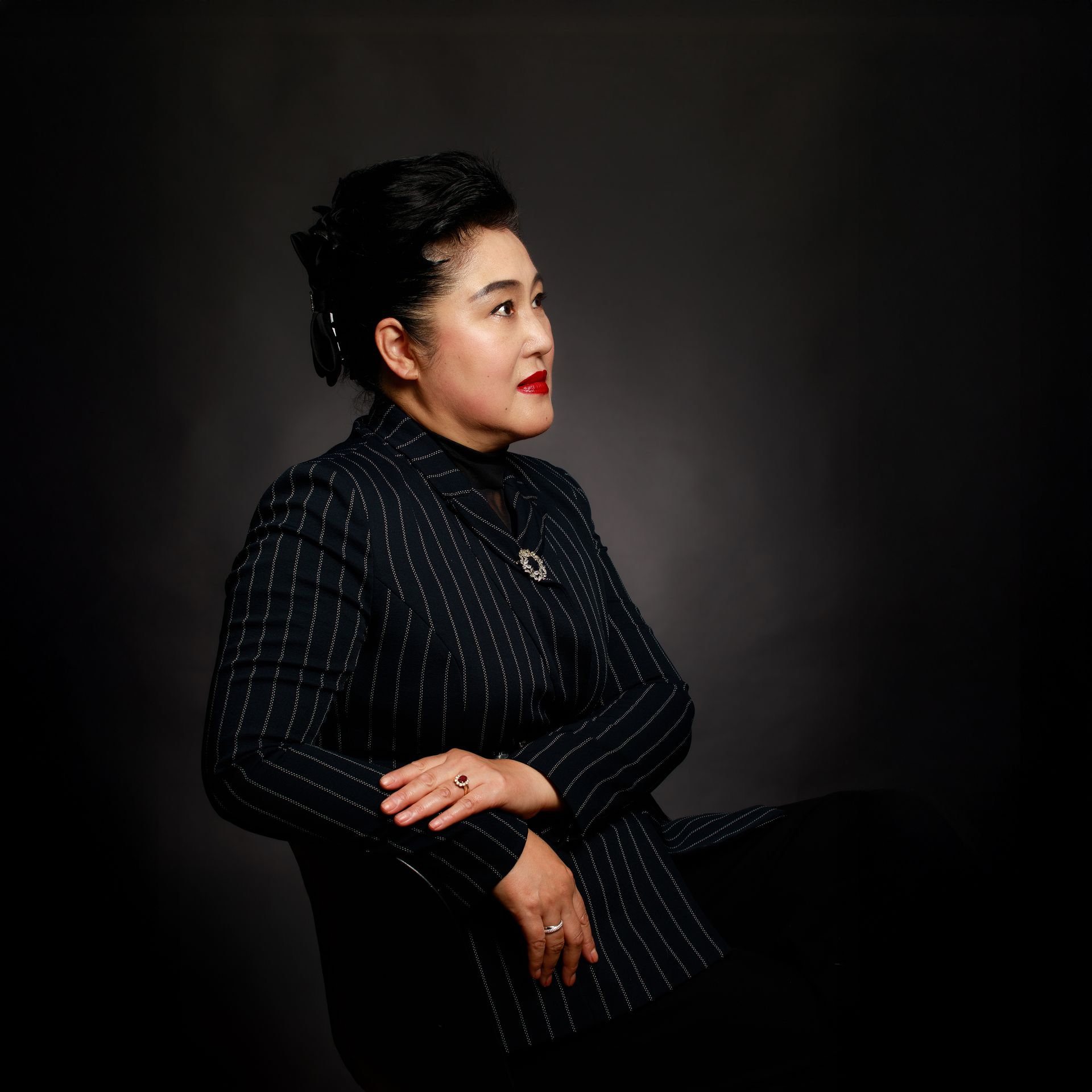 Femme en veste à fines rayures, regardant sur le côté, assise devant un fond sombre