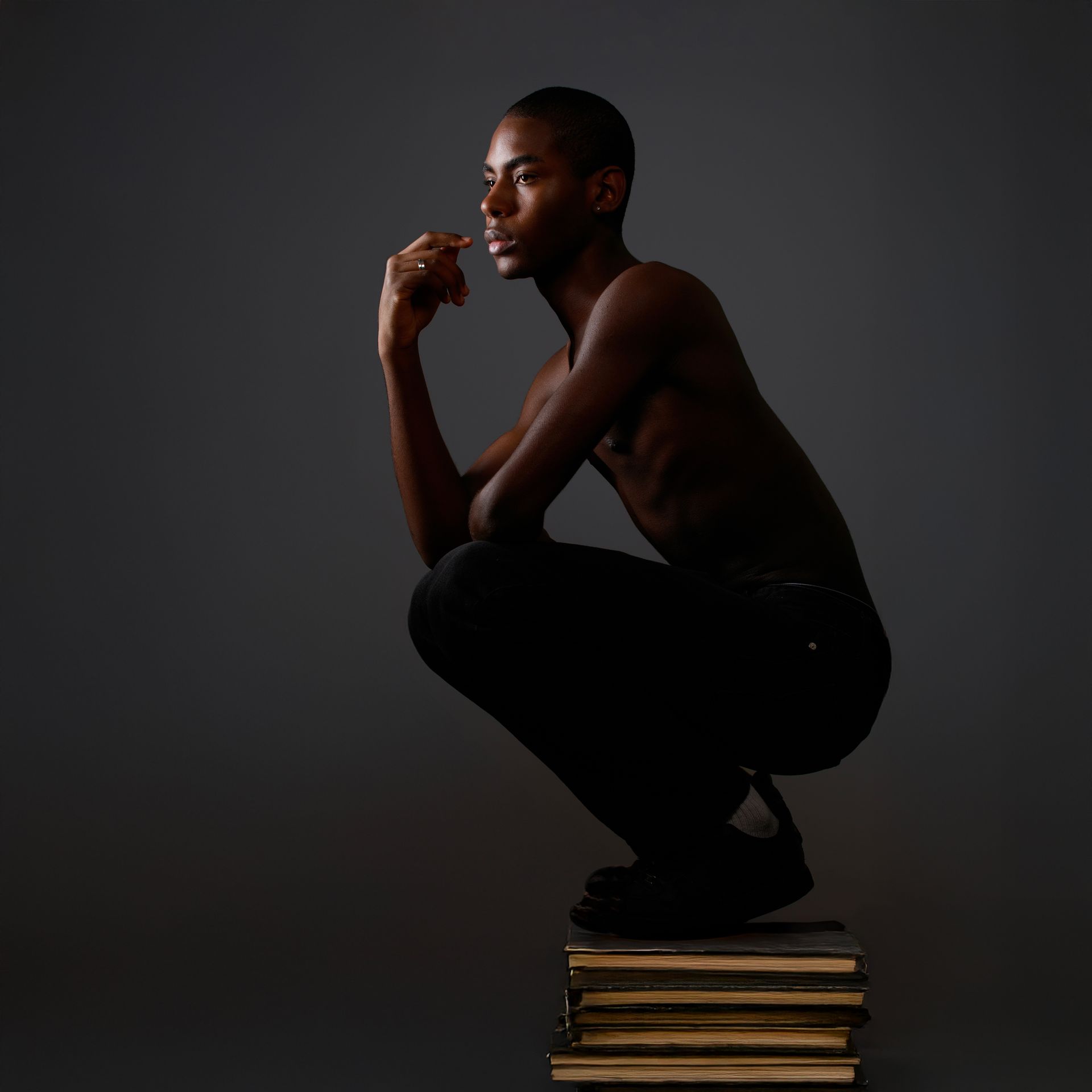 Un homme est accroupi sur des livres, la main sur le menton, le regard détourné