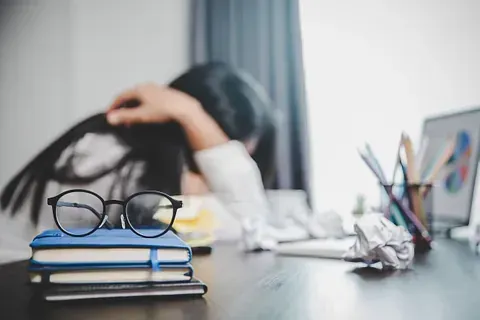 Mujer con la cabeza entre las manos, abrumada en el escritorio con cuadernos, gafas y papel arrugado.