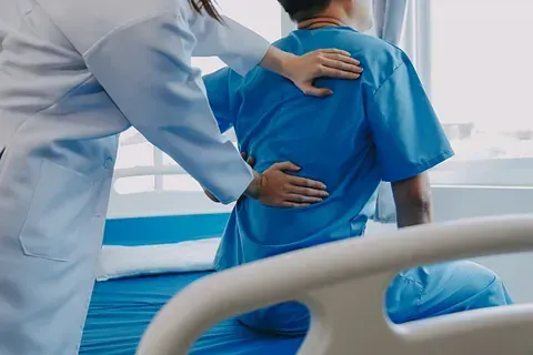 Médico ayuda a un paciente con uniforme azul a sentarse en una cama de hospital.