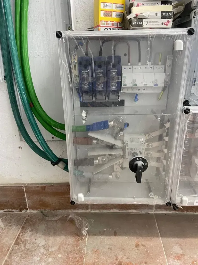 Una caja transparente llena de equipos eléctricos se encuentra sobre un piso de baldosas al lado de una manguera verde.