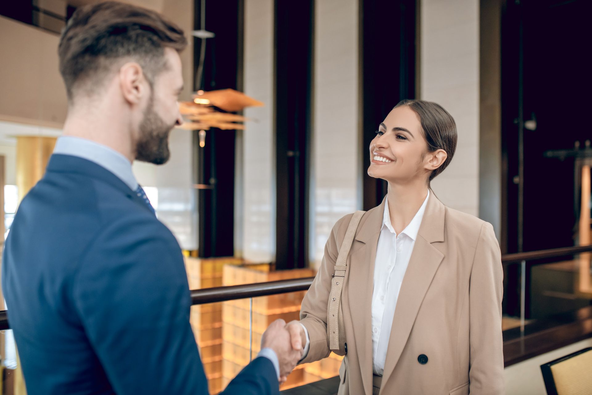 Un hombre con traje azul estrecha la mano de una mujer con blazer color canela, ambos sonriendo. Interior.