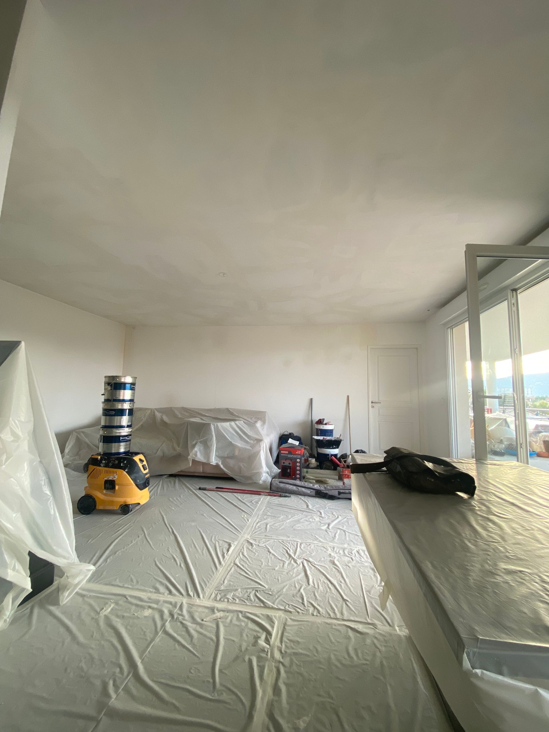 Pièce à rénover avec sol protégé par un  plastique, pots de peinture et aspirateur
