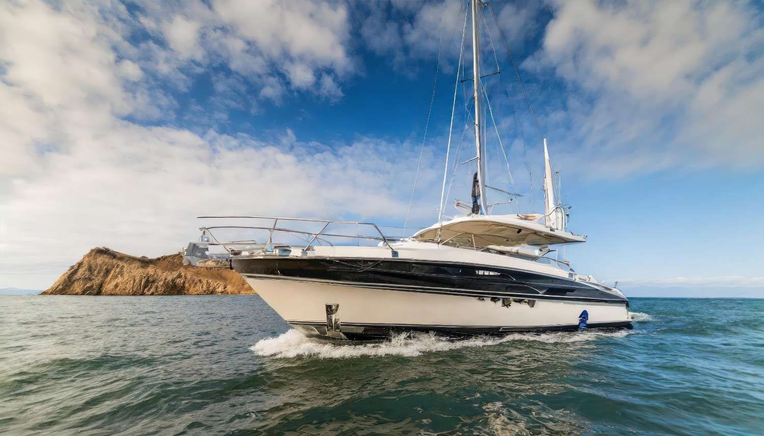 Yate blanco y negro navegando en aguas azules, bajo un cielo parcialmente nublado, con una pequeña isla al fondo.