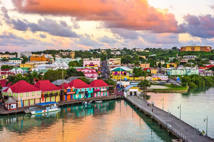 Colorful waterfront town at sunset with red-roofed buildings, a pier, and calm reflective water in St. John's Antigua.
