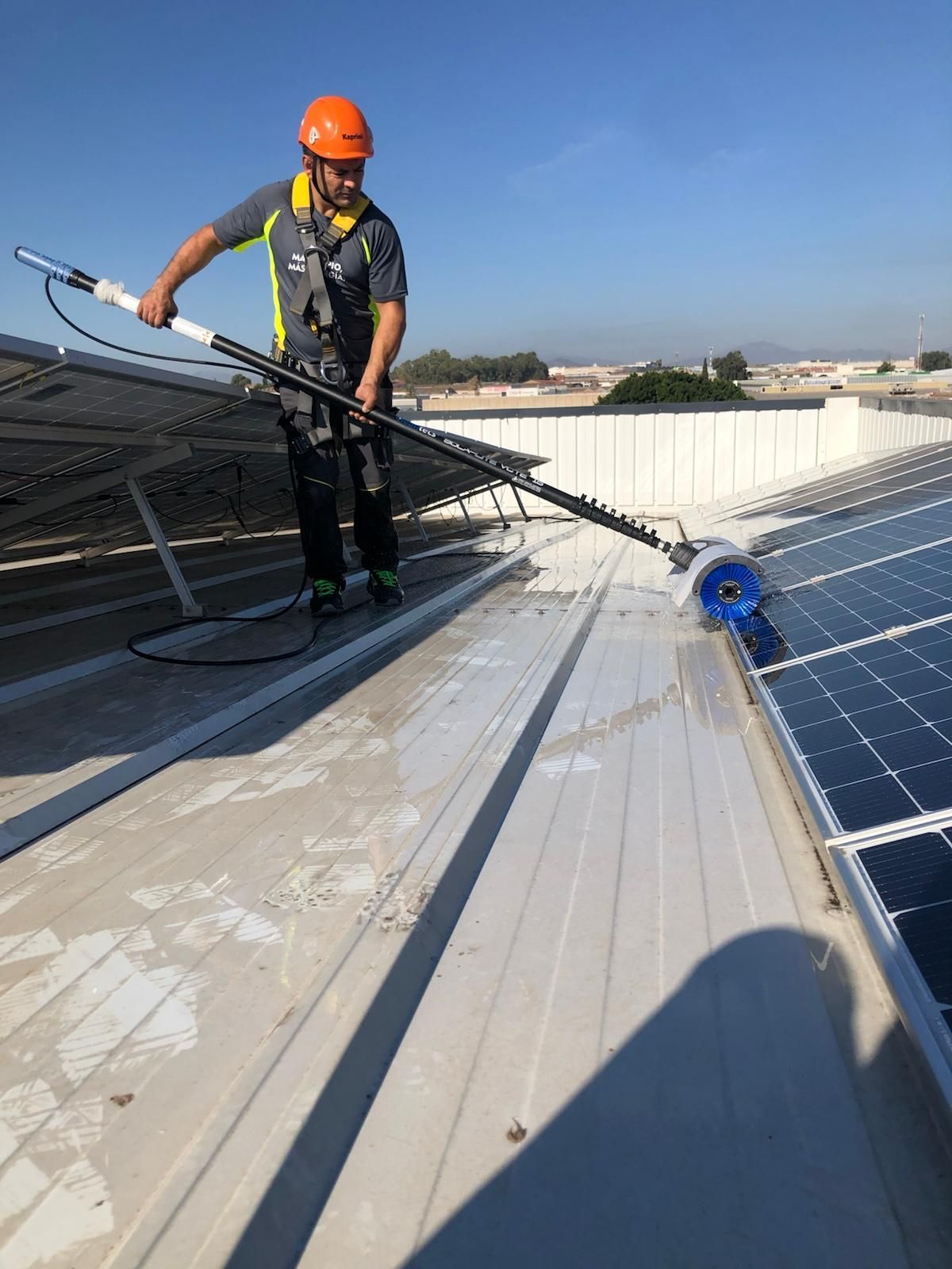 Hombre con arnés limpiando paneles solares en un tejado con un cepillo extensible.