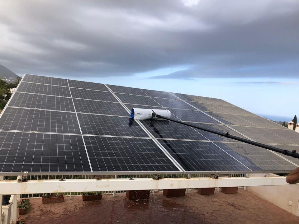 Paneles solares siendo limpiados con un cepillo de mango largo en un tejado con cielo nublado de fondo.