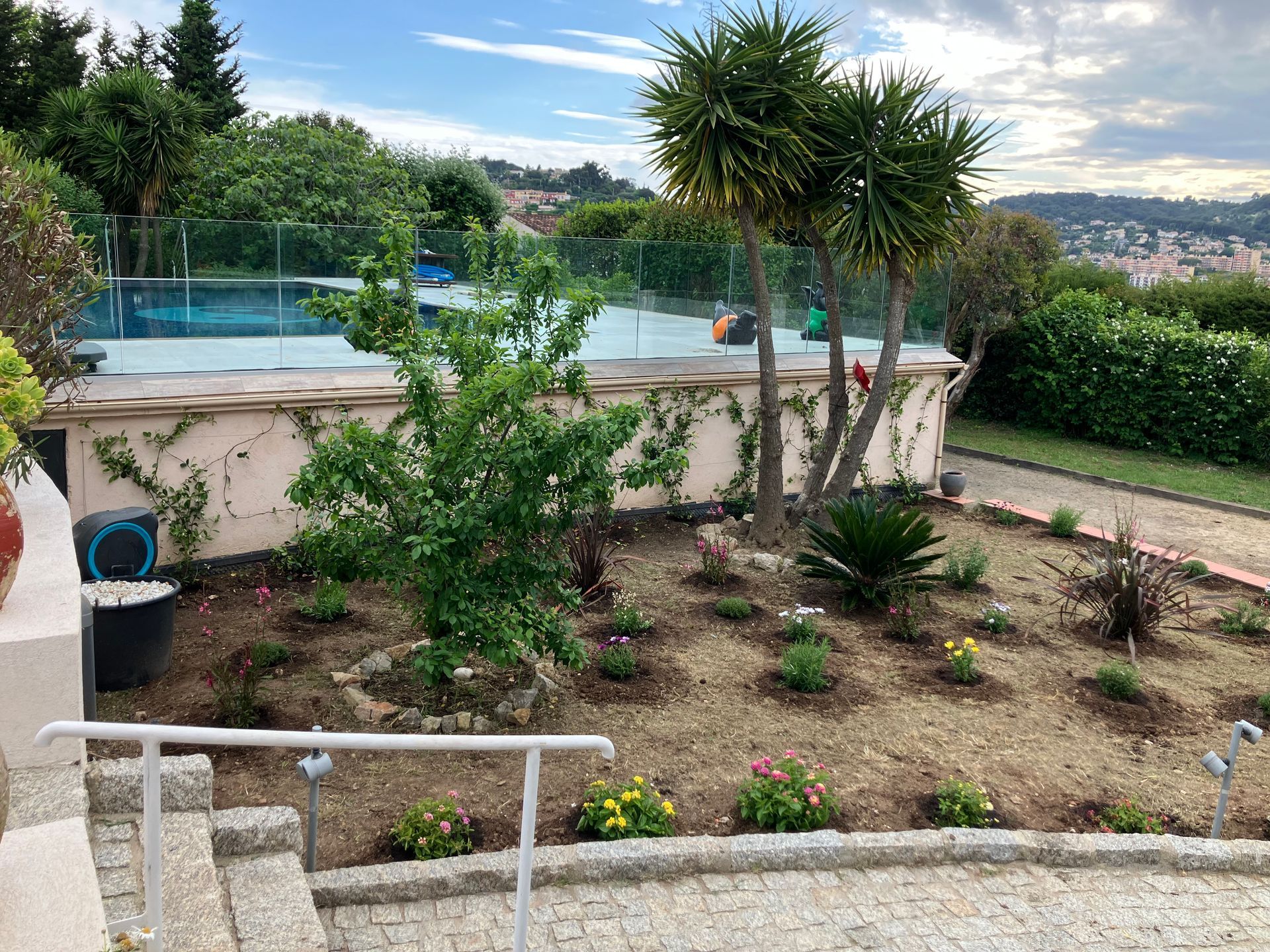 Jardin arrière avec arbustes et petits arbres à côté d'une piscine clôturée sous un ciel nuageux