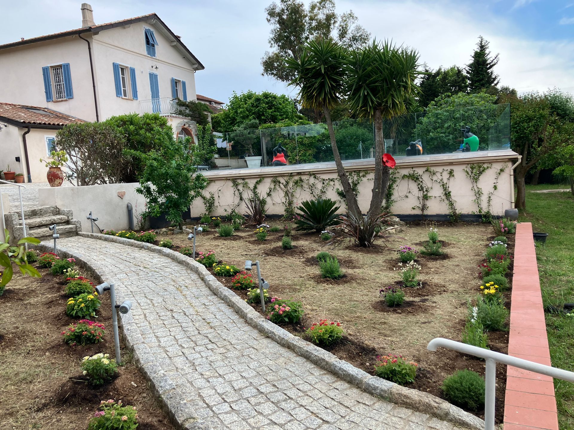 Chemin de pierre sinueux traversant un jardin paysager, à côté d'une maison couleur crème aux volets bleus.