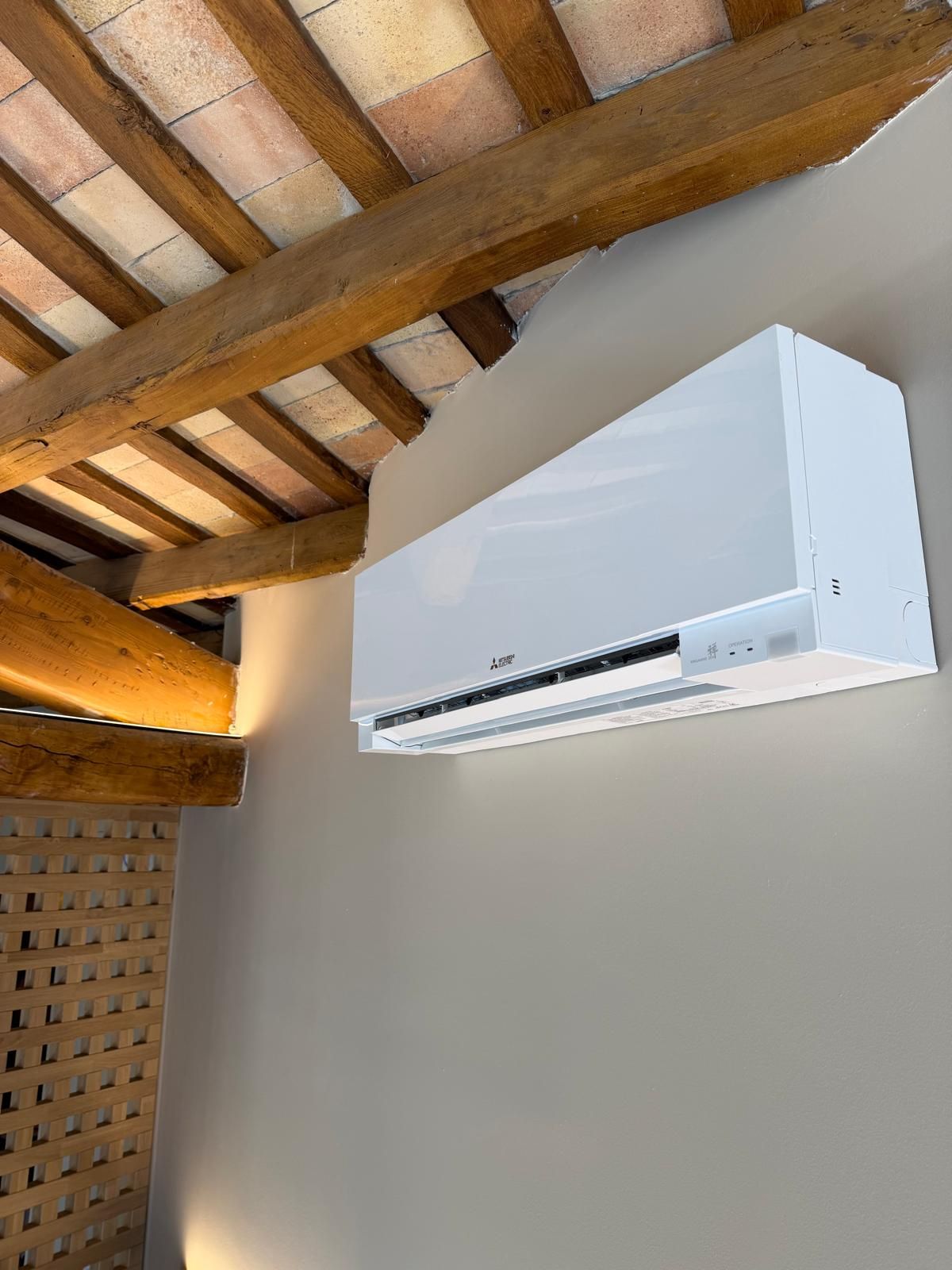 Unidad de aire acondicionado blanca montada en una pared beige, bajo un techo con vigas de madera.