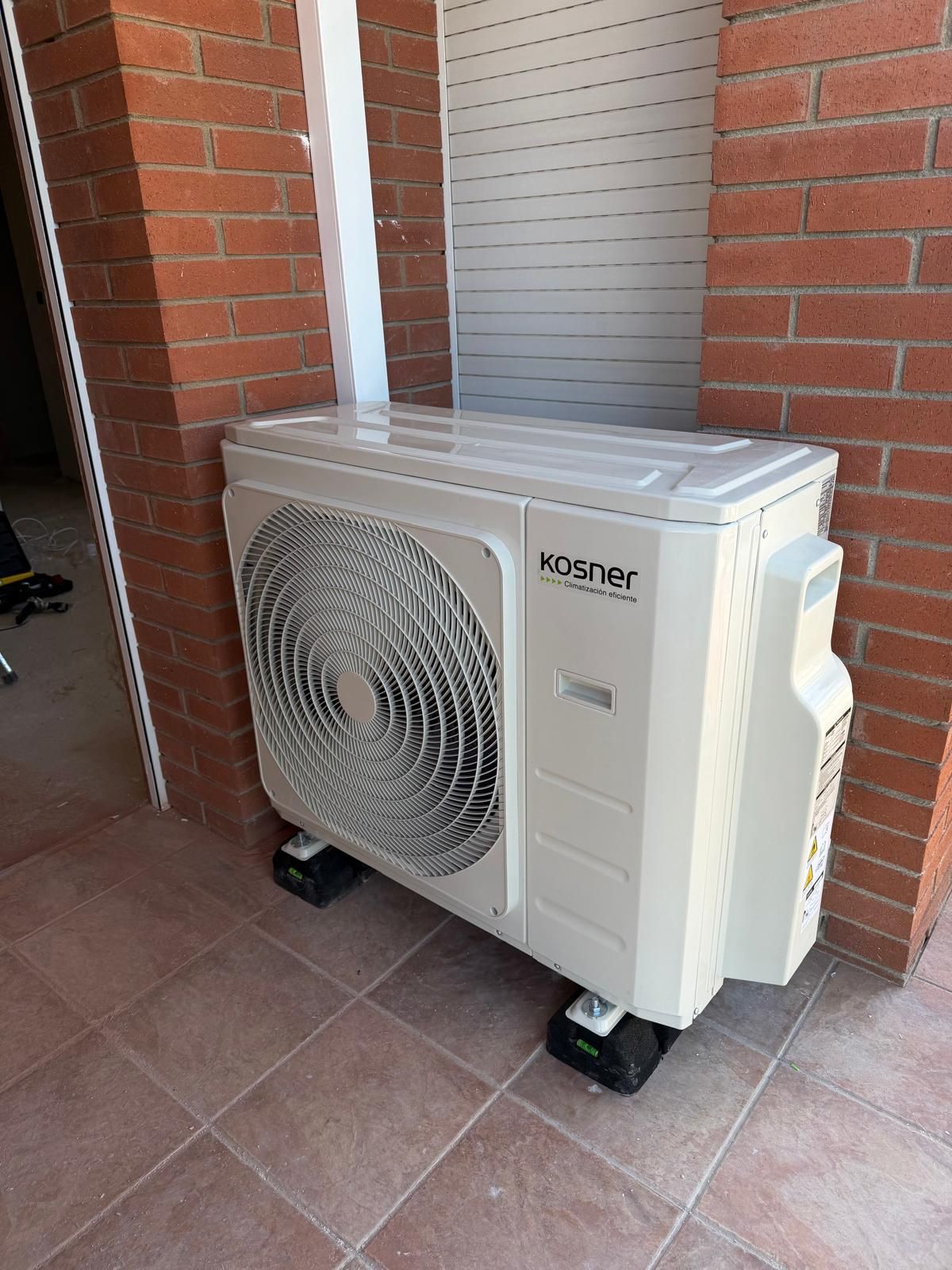 Bomba de calor Kosner blanca y gris sobre soportes negros, junto a una pared de ladrillo y una columna blanca.