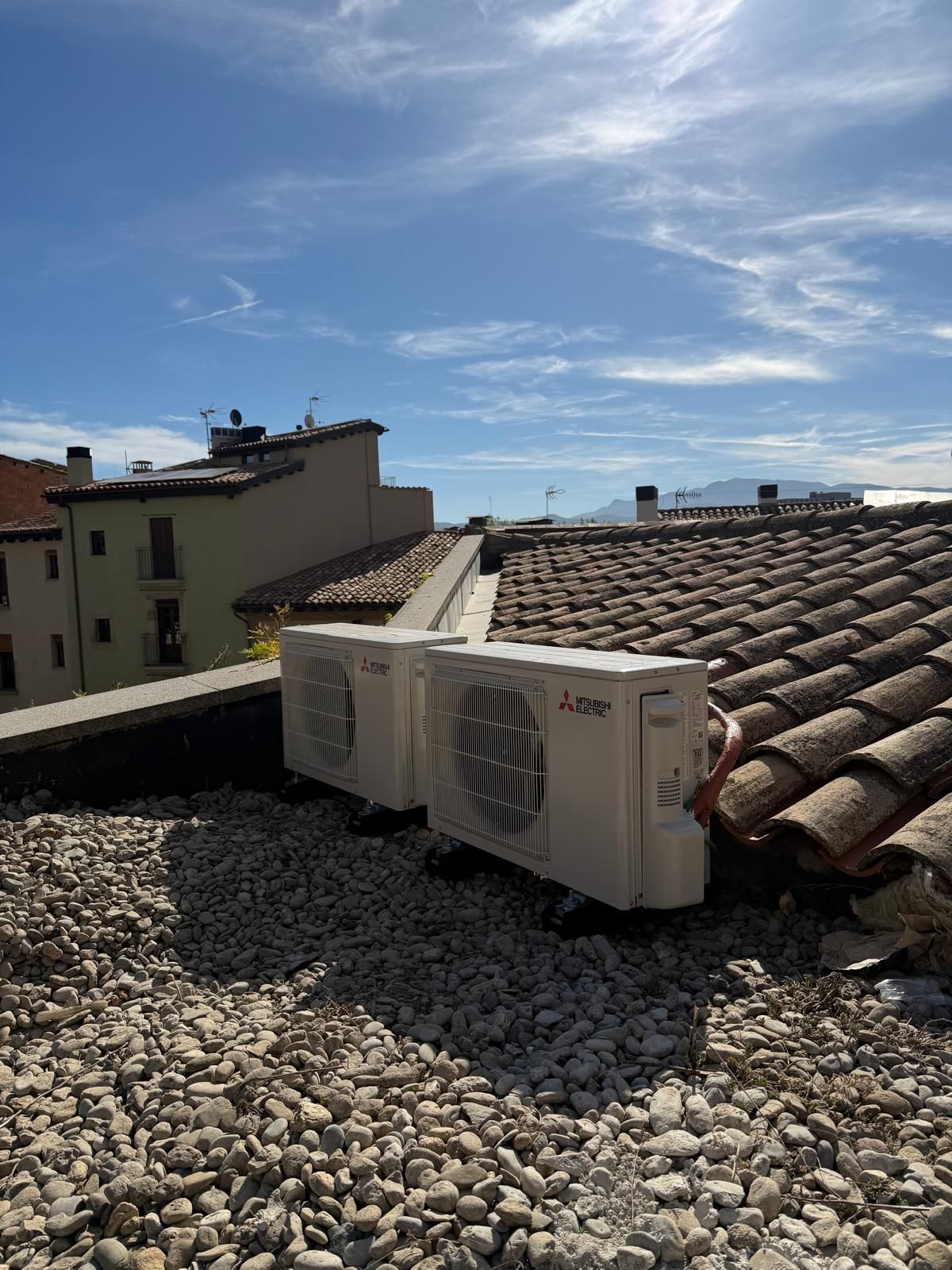 Dos unidades de aire acondicionado blancas en una azotea con tejas de terracota y grava, edificios y cielo al fondo.
