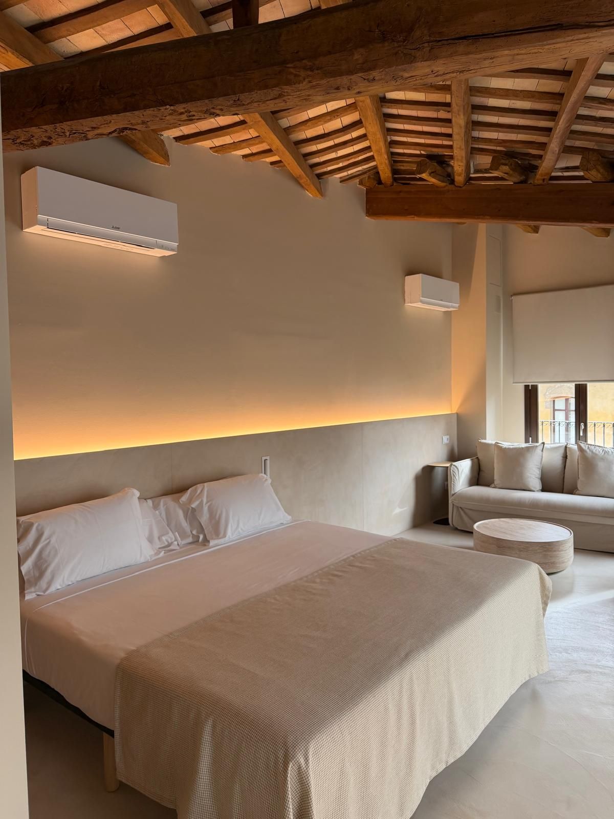 Dormitorio con cama, sofá y unidades de aire acondicionado bajo un techo con vigas de madera.