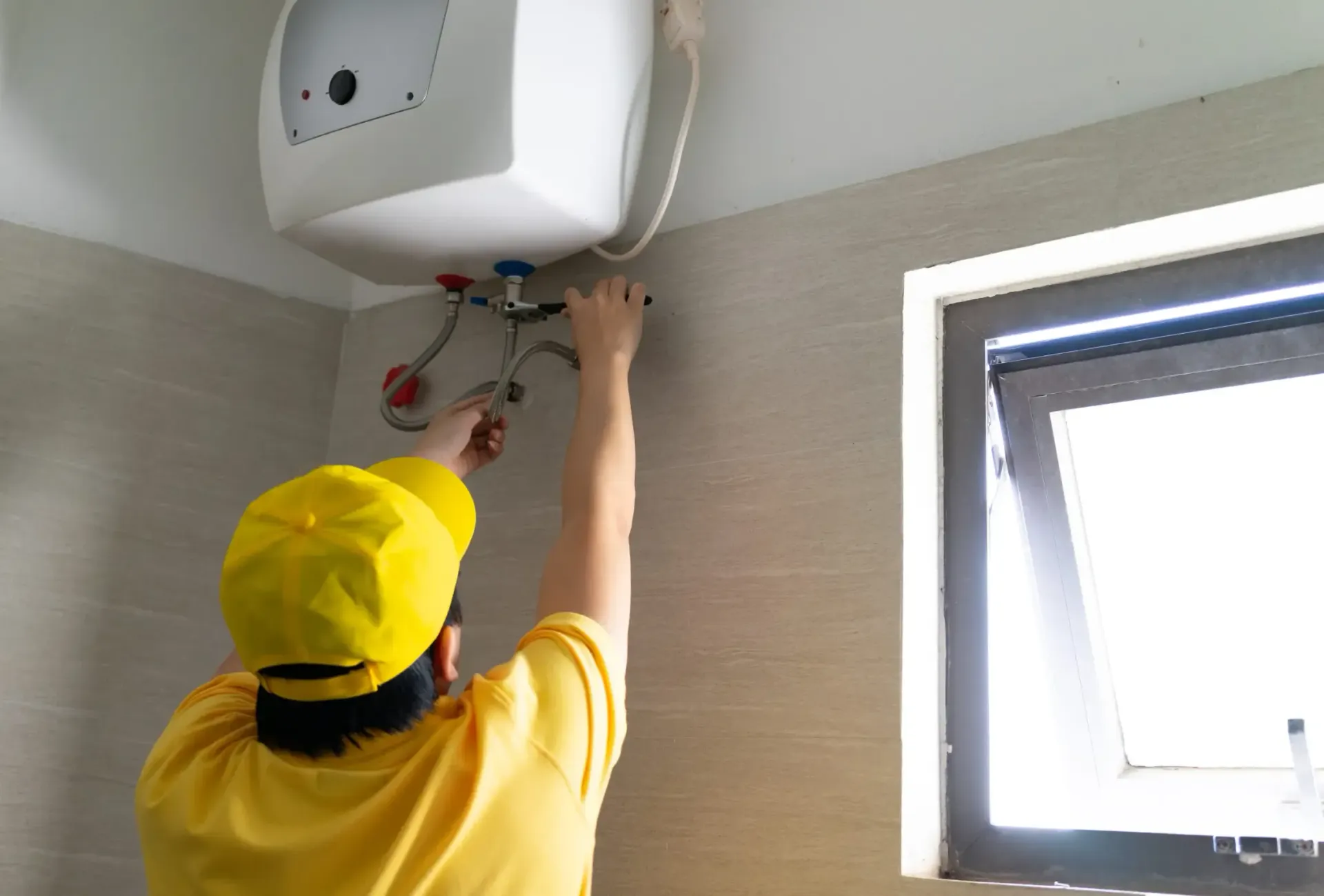 Técnico con camisa amarilla y casco trabajando en un calentador de agua mural cerca de una ventana.