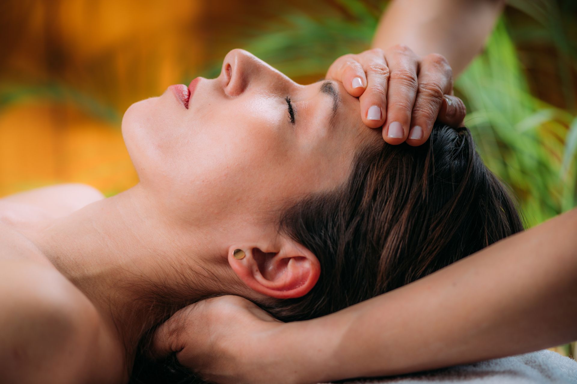 Eine Person, die in einem Spa eine Stirnmassage erhält, liegt zurückgelehnt, hat die Augen geschlossen und die Hände auf dem Kopf ruhen.